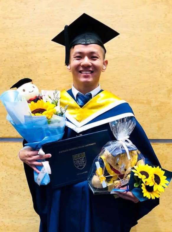 Smiling male graduate in blue and gold gown holding a diploma, sunflowers, and a stuffed bear gift.